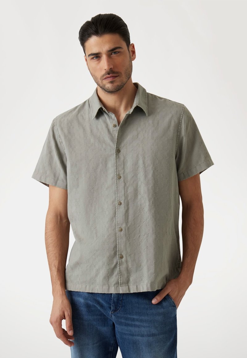 Camisa de manga corta, con botones, en verde claro, que presenta una tela texturizada con patrones sutiles y un cuello estándar. Combinada con jeans azules.