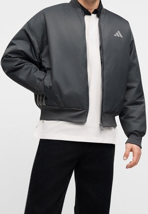 Homme portant une veste bomber gris foncé zippée sur une chemise blanche et un pantalon noir, une main dans la poche de la veste.