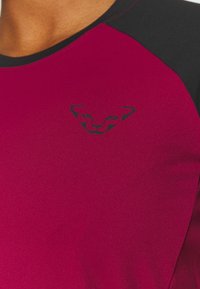 Dopasowana sportowa koszulka w głębokim kolorze magenty z czarnymi akcentami na ramionach. Posiada małe czarne logo na piersi. Gładki, elastyczny materiał.