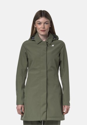 Giovane donna che indossa un impermeabile verde oliva aderente con cappuccio e un piccolo logo bianco sul petto sinistro, in piedi davanti a uno sfondo uniforme.