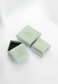 Cajas de papel verde claro con acentos dorados, de tamaños variados, con interior negro, textura suave y branding impreso en las superficies.