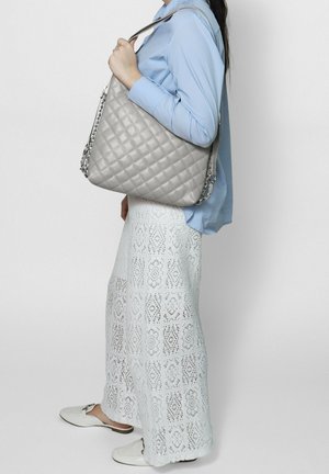 Femme vêtue d'une chemise bleue et d'un pantalon en dentelle blanche, portant un grand sac bandoulière matelassé gris avec une lanière en chaîne.