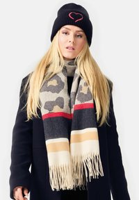Blonde woman wearing black coat, animal print scarf with red, black, beige stripes, and black knit hat with red heart outline.