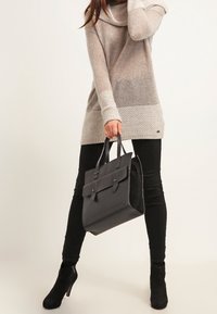 Sac en cuir noir avec deux poignées supérieures, texture lisse, forme rectangulaire et design minimaliste ; associé à un pull en tricot beige et un pantalon noir.