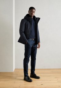 Marine parka-jack met een capuchon, over een marineblauwe trui, gecombineerd met donkerblauwe broek en zwarte veterschoenen op een houten vloer.