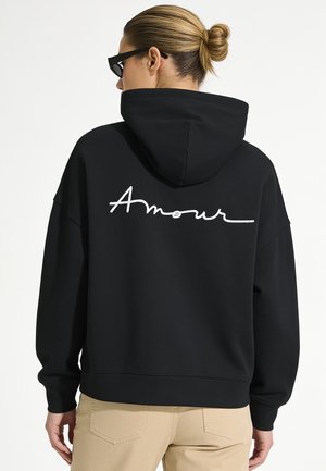Femme avec un chignon portant des lunettes de soleil noires, un sweat à capuche noir avec "Amour" écrit dans le dos, et un pantalon beige sur un fond uni.