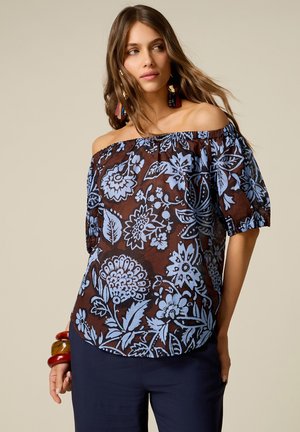 Jeune femme aux longs cheveux porte un haut marron épaules dénudées avec de grands motifs floraux bleus et un pantalon bleu foncé sur un fond uni.