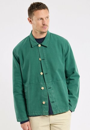 Homme portant une veste verte boutonnée par-dessus une chemise sombre et un pantalon beige, regardant sur le côté sur un fond uni.