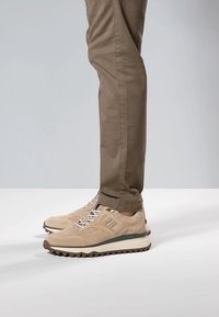 Zapatillas deportivas de ante beige con una parte superior texturizada, entresuela blanca, suela exterior de goma oscura y detalles en gris contrastante. Combinadas con pantalones marrones.