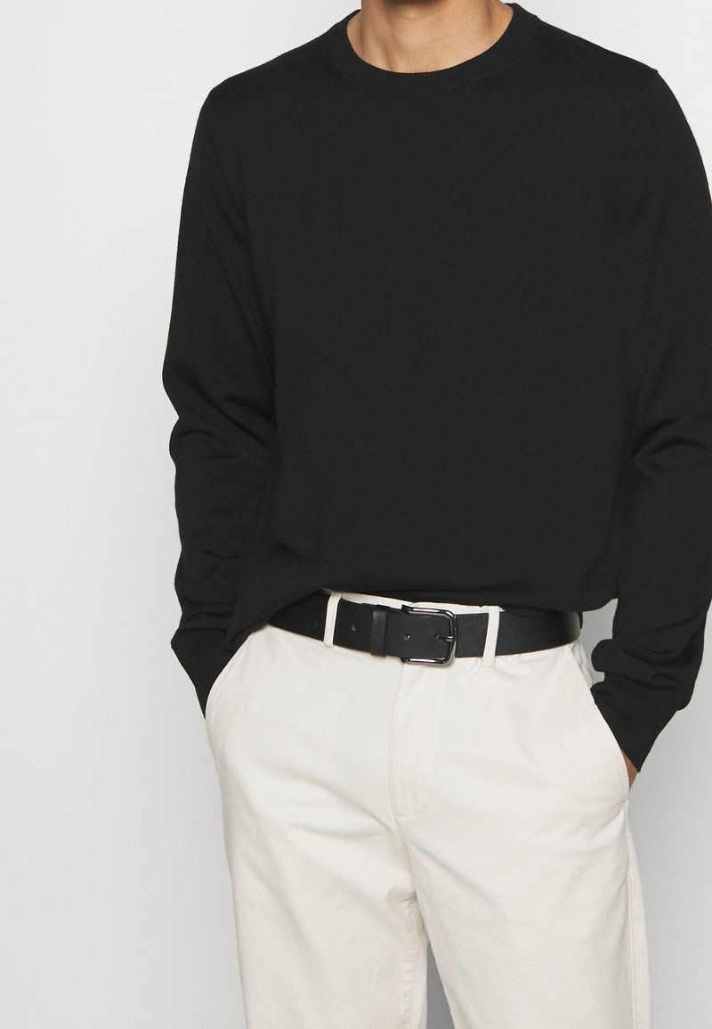 Homme portant une chemise noire à manches longues rentrée dans un pantalon blanc cassé avec une ceinture noire, mains dans les poches, sur un fond clair uni.