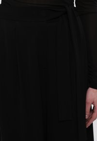 Čierny top z látky s opaskom, ktorý má viazanú stuhu, mierne priesvitné dlhé rukávy a plisované detaily v oblasti pása. Hladká textúra a priliehavý strih.