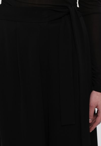 Black fabric belted top featuring a tied sash, slightly sheer long sleeves, and pleated detailing at the waist. Smooth texture and form-fitting design.