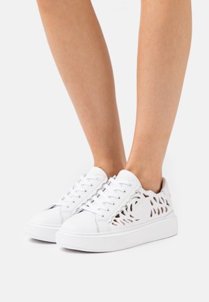 Hvide lavtopsneakers med blomsterudskæringsmønstre, båret på bare fødder, stående mod en ensartet lys baggrund.