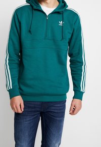 Man som bär en teal Adidas-hoodie med vita ränder på ärmarna och en frontdragkedja, tillsammans med mörkblå jeans, stående mot vit bakgrund.