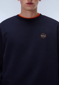 Ciemnogranatowy sweatshirt z okrągłym dekoltem i pomarańczowym wykończeniem wewnętrznym, z okrągłą łatą z logo w górnym lewym rogu. Gładka tekstura.