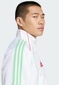 Vit sportjacka med gröna och röda ränder, hög krage, dragkedja och präglad design fram. Adidas-logotyp i rött.