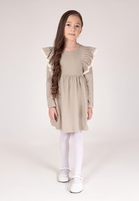 Beżowa sukienka z falbaniastymi rękawami i koronkową lamówką, zebrana w talii, w zestawieniu z białymi rajstopami i butami typu Mary Jane. Gładka tkanina i prosty design.