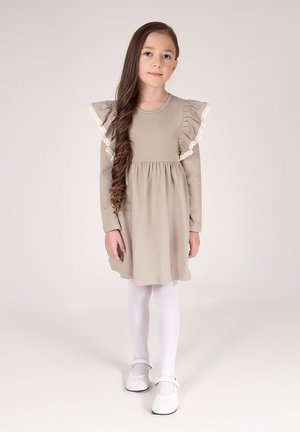 Beżowa sukienka z falbaniastymi rękawami i koronkową lamówką, zebrana w talii, w zestawieniu z białymi rajstopami i butami typu Mary Jane. Gładka tkanina i prosty design.