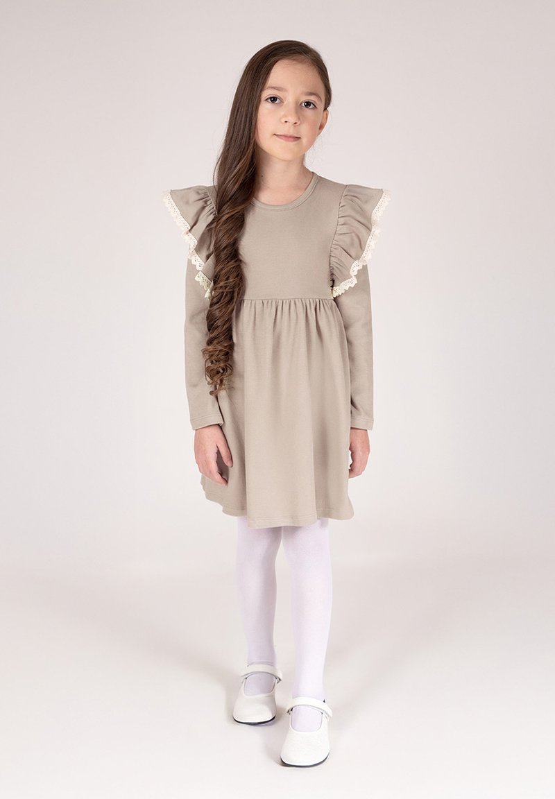 Beżowa sukienka z falbaniastymi rękawami i koronkową lamówką, zebrana w talii, w zestawieniu z białymi rajstopami i butami typu Mary Jane. Gładka tkanina i prosty design.