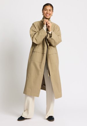 Uśmiechnięta kobieta w beżowym długim płaszczu, kremowych spodniach i czarnych butach, stojąca na tle jednolitego białego tła.