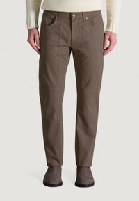 Uomo che indossa pantaloni marroni a gamba dritta con dettagli della cucitura frontale in vista, abbinati a scarpe in suede taupe e un maglione in lana color crema.