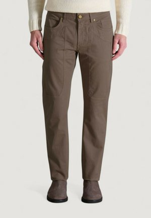 Hombre que lleva pantalones rectos marrones con detalles de costura frontal visibles, combinados con zapatos de ante color topo y un suéter de punto en color crema.
