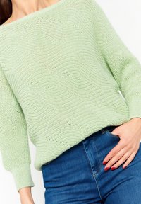 Pull en tricot vert clair avec un large col et un motif texturé en vagues. Manches longues, associé à un jean en denim bleu foncé.