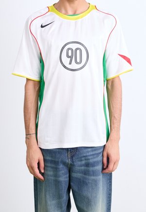 Homme portant un maillot de sport blanc avec le numéro 90 dans un cercle, col jaune, liseré rouge, panneaux latéraux verts et jean bleu ample.