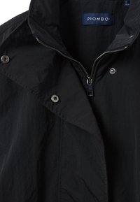 Chaqueta ligera negra con un alto cuello, cierre de cremallera y botones a presión. Presenta una tela ligeramente texturizada y un diseño minimalista.