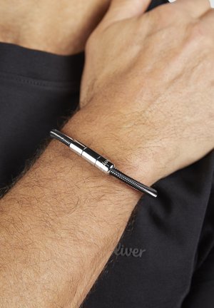 Black braided bracelet with silver hardware and a cylindrical clasp, worn on a wrist with light brown skin, against a dark shirt background.