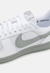 Vita sneaker i slät läder med grå detaljer, texturerad gummisulor och sex vita snörhål; har en framträdande Nike-logotyp.