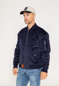 Veste bomber bleu marine avec une texture lisse et douce, des poignets côtelés, un zip sur le devant et une étiquette orange sur l'ourlet. Comprend des poches latérales.