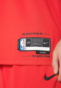 Camiseta de la NBA roja con textura de malla. Presenta el logo negro de Nike, una etiqueta con "Diseñada según las especificaciones exactas" y talla 48.