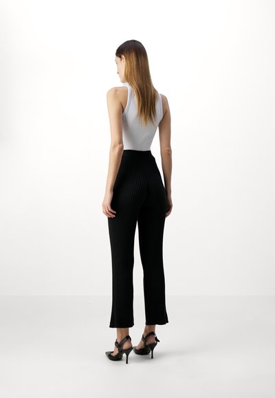 Black ribbed pants with a flared hem, paired with a fitted gray tank top. Model wearing black high-heeled sandals, seen from the back.