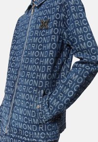 Giacca in denim con tessuto blu caratterizzato da un motivo ripetuto con la scritta "RICHMOND", chiusura con zip e tasche laterali. Accentuata da un badge con logo.