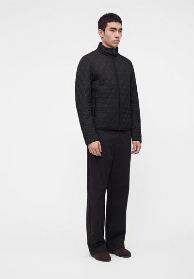 Homme portant une veste matelassée noire avec des poches zippées et un pantalon noir, debout devant un fond blanc uni.