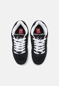 Sorte joggesko laget av suede og tekstil, med hvite snørebånd, polstret krage og en rød logopatch på tungen, med et klassisk design.