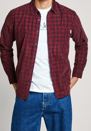 Mand iført en åben rød ternet skjorte over en hvid grafisk t-shirt, kombineret med blå denimjeans, hænderne i lommerne.