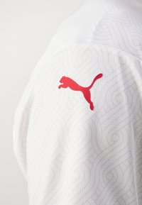 T-shirt de sport blanc avec un motif texturé ; présente un logo Puma rouge sur la manche, mettant en valeur son design en tissu respirant.