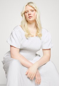 Vestido branco com corpete franzido e mangas bufantes, apresentando um tecido texturizado. O modelo tem cabelo loiro longo e ondulado e está sentado em um fundo neutro.