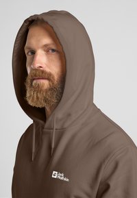 Felpa marrone realizzata in tessuto morbido, con cappuccio regolabile e un piccolo logo bianco Jack Wolfskin sulla parte sinistra del petto.