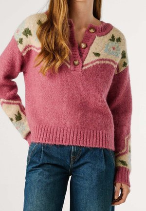 Femme portant un pull en maille rose avec un empiècement crème à motif floral et des boutons en bois, associé à un jean en denim bleu.