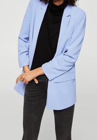 Personne portant un blazer bleu clair sur un haut noir et un jean foncé, debout devant un fond clair uni, les bras croisés.
