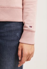 Handen avslappnad bredvid mörkblå jeans, med en ljusrosa ribbad sweatshirt med en liten röd, vit och blå logotyp nära handleden.