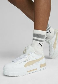 Zapatillas deportivas de plataforma blancas con acentos en beige claro, de cuero suave y malla texturizada. Combinadas con calcetines blancos que tienen rayas negras.