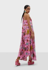Robe maxi en tissu rose, ornée de motifs floraux de différentes couleurs. Présente des bretelles fines et une silhouette fluide, accompagnée de sandales à talons compensés roses.
