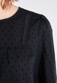 Black blouse featuring textured polka dots, lace detailing along the shoulder seam, and a round neckline. Soft, lightweight fabric.