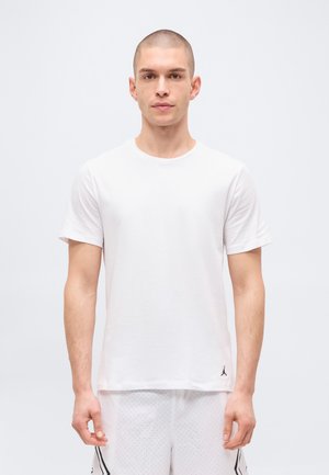 Άνδρας μοντέλο με κοντά μαλλιά που φορά απλό λευκό κοντομάνικο μπλουζάκι και λευκά αθλητικά σορτς με μικρές μαύρες κουκκίδες.