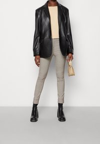 Personne portant un blazer en cuir noir, un haut beige, un pantalon slim à carreaux, des bottes à plateforme noires, tenant un petit sac à main beige sur fond blanc.