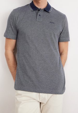 Mann trägt ein kurzärmeliges graues Polohemd mit marineblauem Kragen und zwei Knöpfen, kombiniert mit hellen Hosen vor einfachem Hintergrund.